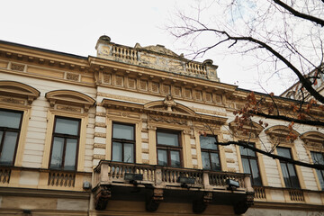 Fototapeta premium Historic building facade with decorative balcony and classical ornaments in Belgrade old town, framed by bare tree branches on a cloudy day.