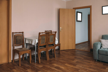 Fototapeta premium A cozy hotel room with wooden flooring. A small dining table with four chairs is set against a light pink wall. A door leads to another room, and a sofa is visible.