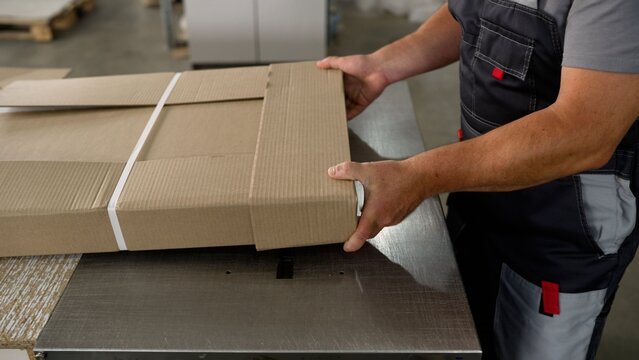 Industrial worker operating a semi-automatic strapping machine to secure a cardboard package with plastic bands in a factory warehouse