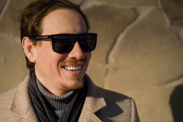Obraz premium Close-up portrait of a young caucasian redhead man with sunglasses smiling wearing a coat on a winter day while walking down the street. Lifestyle.