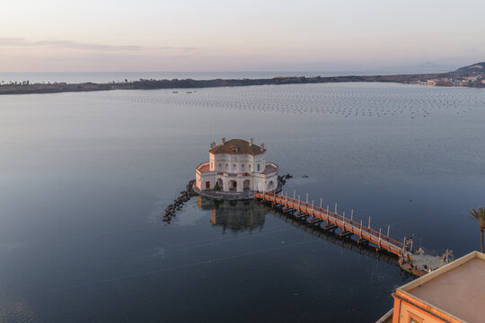 Aerial view of Casina Vanvitelliana's elegant architecture mirrored in the Fusaro Lake's tranquil waters under a soft sky, Bacoli, Campania, Italy.