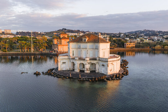 Aerial view of the Casina Vanvitelliana, a delicate palace rising from the tranquil waters of Fusaro Lake, its pale facade reflecting the soft, golden light of dawn, Bacoli, Campania, Italy.