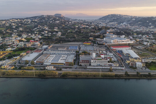 Aerial view of industrial buildings meeting the calm waters of a lake under a sky of soft hues, Fusaro Lake, Bacoli, Campania, Italy.