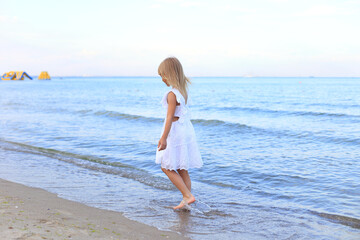 A young girl in a white dress walks along the shoreline, her bare feet splashing in the gentle waves under a bright, clear sky.
