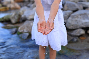 A child in a white dress stands in a rocky stream, gently cupping water in her hands as droplets fall back into the clear, flowing current.