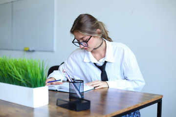 A focused young woman in a school uniform, complete with a white shirt and black tie, is diligently studying at a wooden desk. She wears glasses and is writing in a notebook