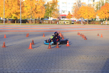 A young boy with dark hair races a go-kart through a course marked by orange cones. Autumn trees with yellow leaves are in the background.