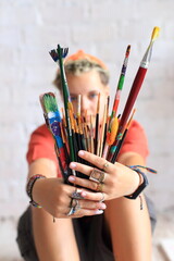 A young artist holds a colorful bundle of paintbrushes toward the camera, their face softly blurred behind the tools of their craft against a white brick wall.