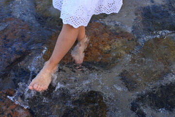 A child in a white dress walks barefoot through a shallow, clear stream, water splashing around her ankles as she steps on smooth, wet rocks.
