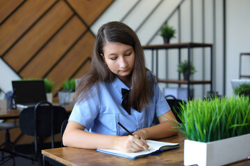 A young student in a light blue uniform concentrates intently while writing in a notebook at a wooden desk. Surrounded by green plants and modern decor