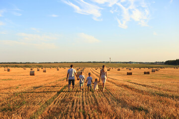 A family walks hand in hand through a golden wheat field during sunset. The scene captures a warm, peaceful atmosphere with hay bales scattered around.