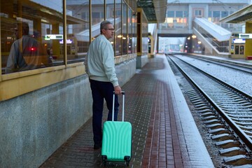 A man stands alone on a snowy, wet platform, holding his bright green suitcase. He looks back over his shoulder as snow dusts the tracks and station, capturing a quiet, solitary moment of travel.