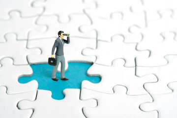 Miniature tiny people toy figure photography. Businessmen standing above missing piece of puzzle jigsaw using binocular