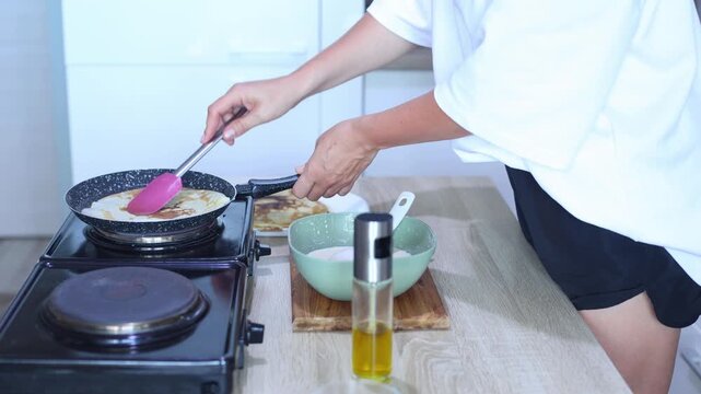 A person uses a pink spatula to carefully lift and flip a pancake in a hot skillet, revealing its perfectly golden-brown cooked side.