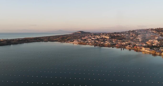 Aerial view of the calm Fusaro Lake reflecting the soft light of dawn amidst coastal buildings and distant mountains, Fusaro Lake, Campania, Italy.