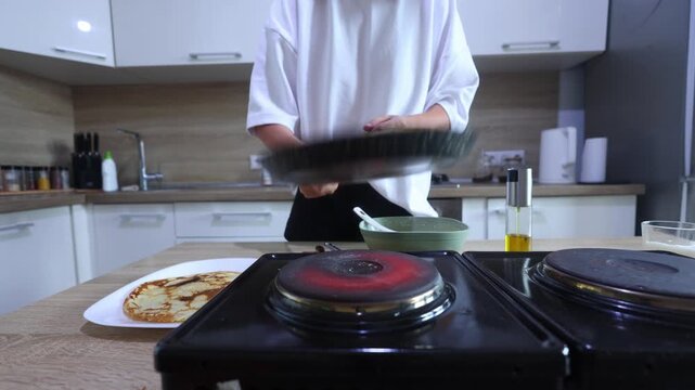 A person flips a golden-brown pancake in a skillet using a flick of the wrist, then slides it onto a plate with another finished pancake, ready to serve.