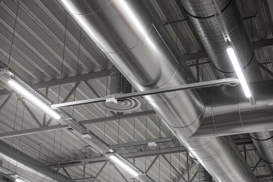 Detailed view of metallic HVAC ducts and ceiling-mounted vents in an industrial interior, featuring suspended LED lighting and steel beams. Clean, modern look with copy space