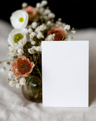 Elegant wedding card mockup displayed next to a beautiful floral arrangement in soft colors on a textured surface during a bright indoor setting