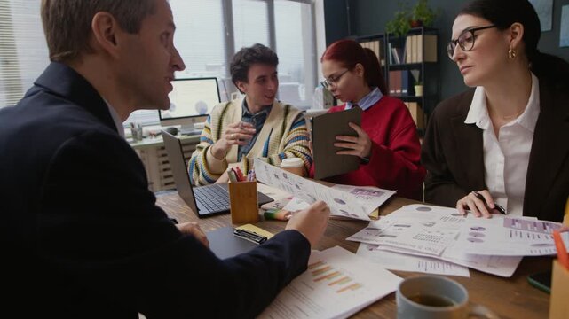 Medium shot of Caucasian colleagues coworking while sitting at office desk with business papers and digital devices