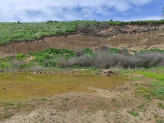 Small summer pond in front of sandy hills with green vegetation, blue sky with clouds and bird nesting holes