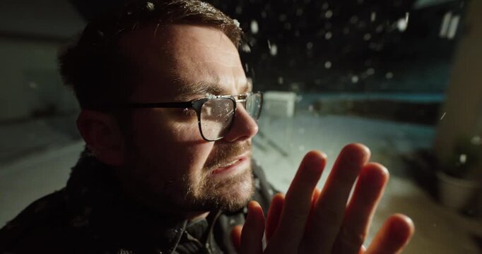 Close-up of a man standing outdoors at night while heavy snow falls. He blows into his hands and rubs them to stay warm. Concept of cold weather, winter discomfort, waiting outside, and human vulnerab