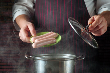 A person in a striped apron holds a plate of sausages over a pot on a stove. Steam rises from the pot while the lid is about to be placed on top