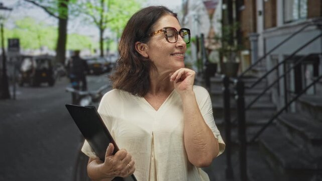 Woman holding folder touches chin on street, wearing glasses and beige blouse with bicycles and stoop visible; thoughtful optimism.
