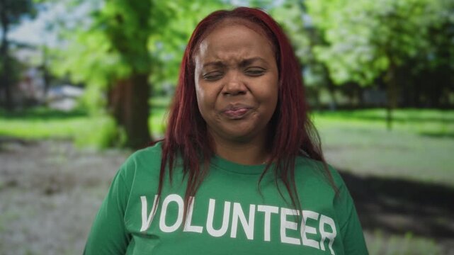 Woman grimaces in green volunteer shirt with visible logo under bright sunlight among tall forest trees; disgust.