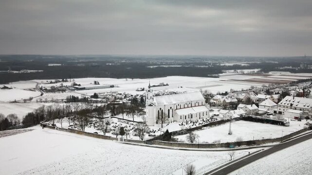 Forward drone flight toward snowy Elchingen church amid village homes, cemetery, snowy fields, roads, hills. Perfect for travel docs, ads, reveals.