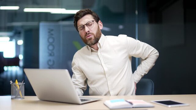Tired businessman sitting in office suffers from back pain. Male employee massages his sore lower back muscles, feeling exhausted and overworked, dealing with tension discomfort from prolonged sitting