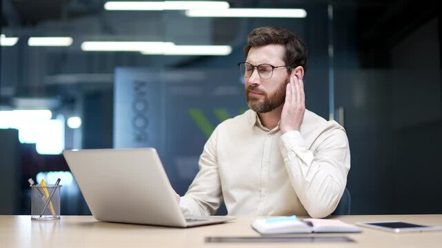Businessman sitting in modern office suffering from ear pain. Male employee touching his ear with fingers, feeling discomfort from infection, otitis media, eczema, or other ear diseases. Earache