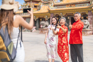 Obraz premium Happy female tourist taking photo of asian family wearing cheongsam at Chinese temple, Senior parents and daughter making mini heart sign, Lunar New Year travel and smartphone technology concept