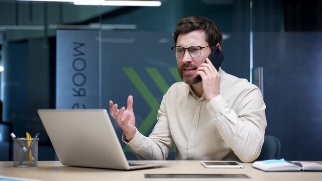 Angry businessman arguing on smartphone sitting at workplace in office. Frustrated manager shouting and gesturing during phone call, expressing stress, anger, and disagreement about business problem