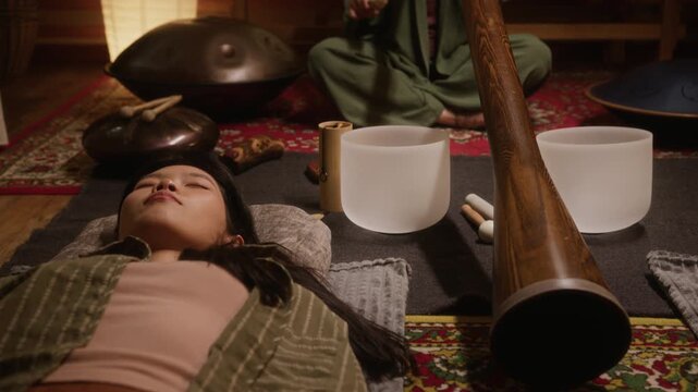 Medium full shot of young Asian girl lying in shavasana on carpet with closed eyes in yoga studio, meditating, practicing mindful breathing at sound healing therapy with didgeridoo and singing bowls