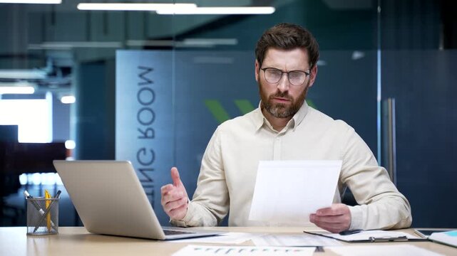 Frustrated puzzled businessman sitting at desk in office, having difficulty with paperwork and laptop. Upset financier reviewing documents, trying to solve a problem, confused and disappointed at work