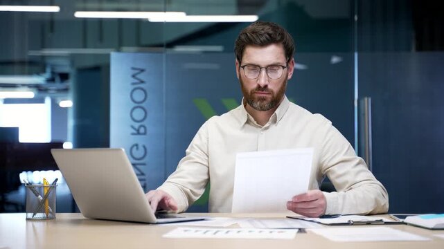Busy businessman sitting at desk in modern office working on laptop and checking documents. Focused financial specialist reviewing reports, analyzing data, and managing company accounting paperwork.