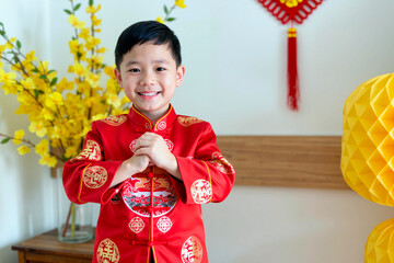 Obraz premium Portrait of Asian boy child smiling and performing traditional greeting gesture while standing indoors during festive celebration, yellow flowers and decorations in background