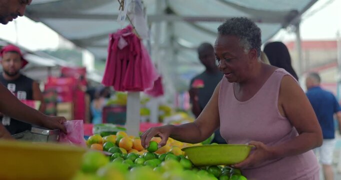 Hispanic street endor holding plastic bag while preparing produce as camera pans toward African American older woman browsing fruit at outdoor market