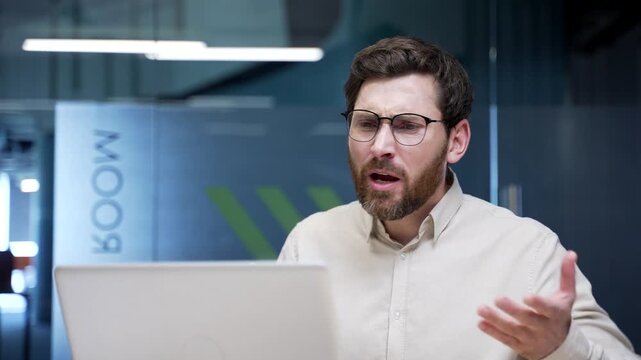 Close-up of angry businessman arguing during video call on laptop in modern office. Frustrated boss talking loudly and gesturing, expressing stress and disagreement while discussing conflict online