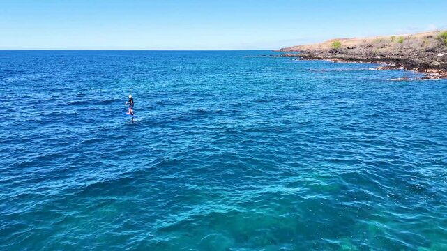 Aerial view of a person hydrofoil surfing over the clear, blue ocean waters under a bright sky, creating a mesmerizing scene, Hawi, Hawaii, United States.