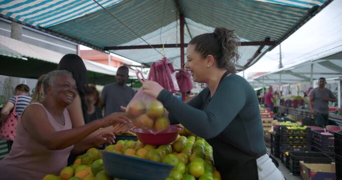 Female street vendor shaking hands with elderly African American customer while holding bag of citrus fruit at outdoor market expressing trust warmth and local food community