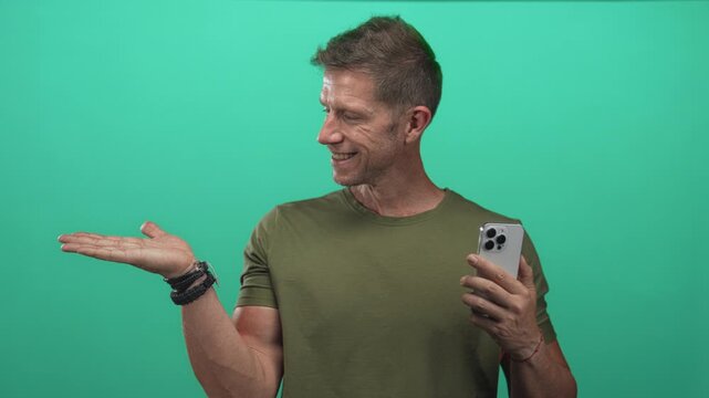 Man holding smartphone and presenting open palm toward camera in green studio; friendly product demo tutorial.