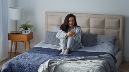 Fototapeta na wymiar Happy Caucasian woman using mobile phone sitting on bed in morning winning with smartphone carefree female girl joyful win victory with online app cellphone in bedroom reading good news notification