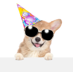 Happy Corgi puppy wearing party cap and sunglasses looking above blank white banner. isolated on...