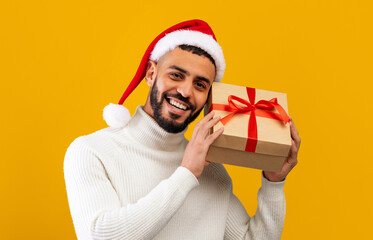 Obraz premium A man wears a red and white Santa hat while smiling and holding a gift box tied with a red ribbon. The background is bright yellow, creating a festive mood for a celebration.