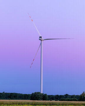 View of a solitary wind turbine standing tall against a gradient sky of lavender and blue, overseeing a field of green and gold, Liw, Masovian Voivodeship, Poland.