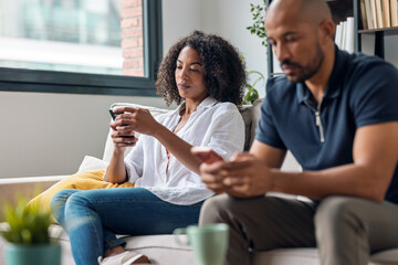 Angry couple sitting on couch together and looking to opposite sides while using smartphone at home.