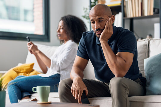 Angry couple sitting on couch together and looking to opposite sides while using smartphone at home.