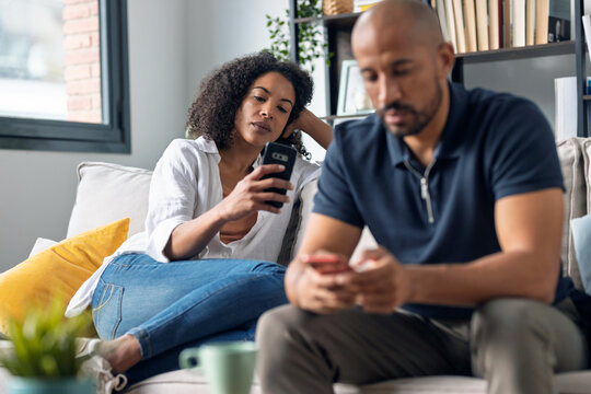 Angry couple sitting on couch together and looking to opposite sides while using smartphone at home.
