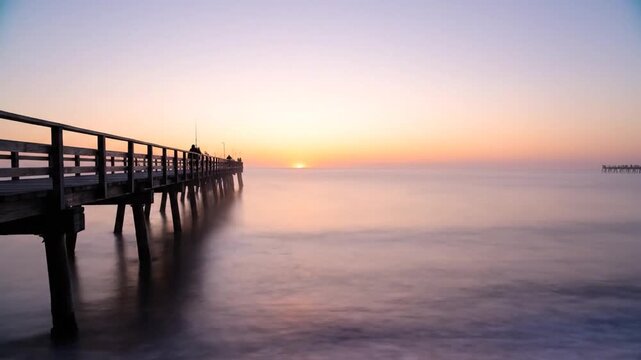 A tranquil time-lapse captures the sun ascending above the horizon over a misty sea. Silhouettes of people stroll and fish along a long wooden pier as the sky shifts from soft violet to brilliant gol
